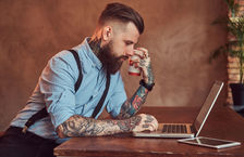 Tattooed hipster working on laptop holding coffee cup