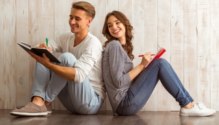 Beautiful young couple writing in a notebook sitting on wooden floor at home